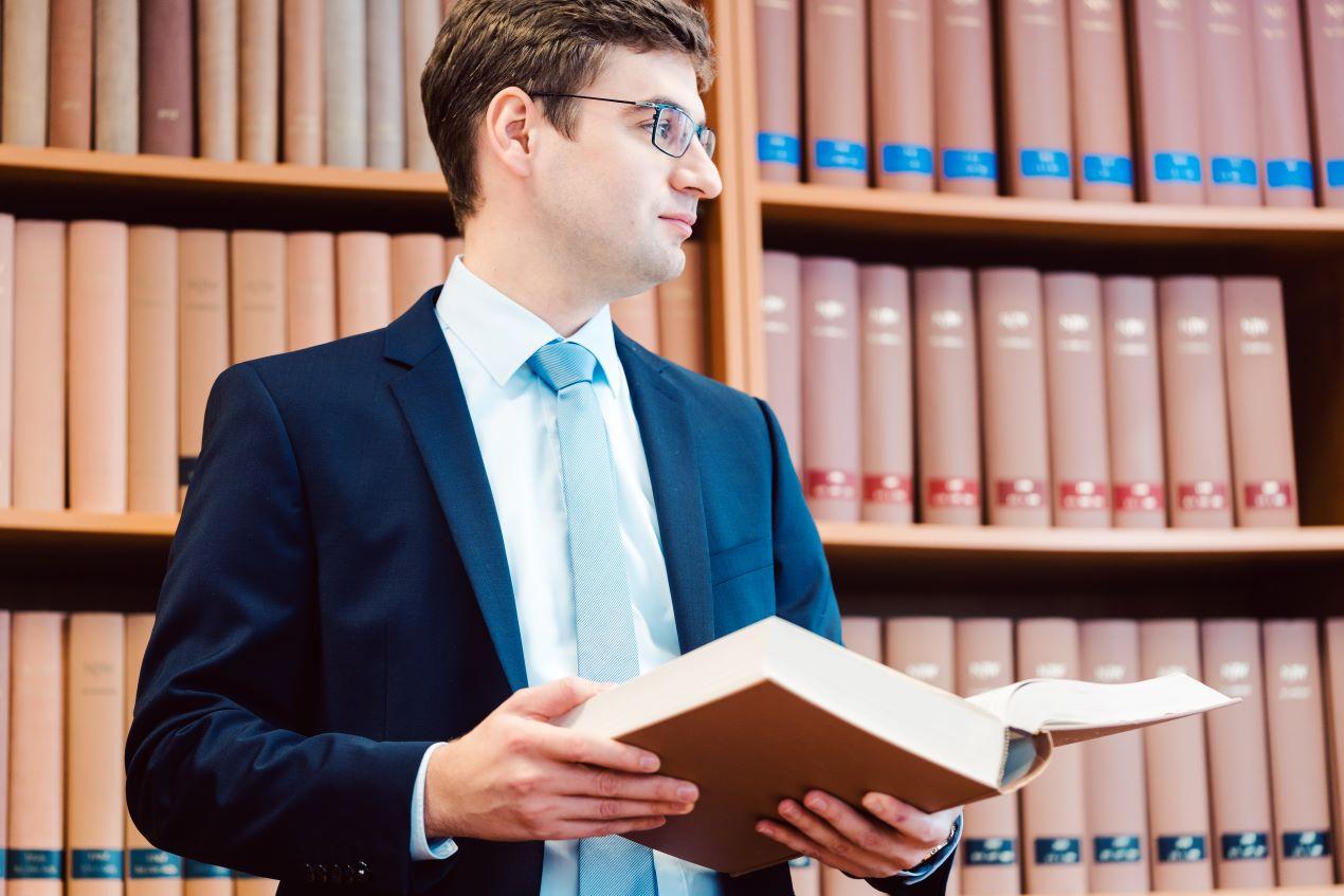 Ein junger Anwalt im Anzug mit einem juristischen Buch in der Hand vor einer Bibliothek. Verdeutlicht die fachliche Expertise der Kanzlei Ra-Hartung.