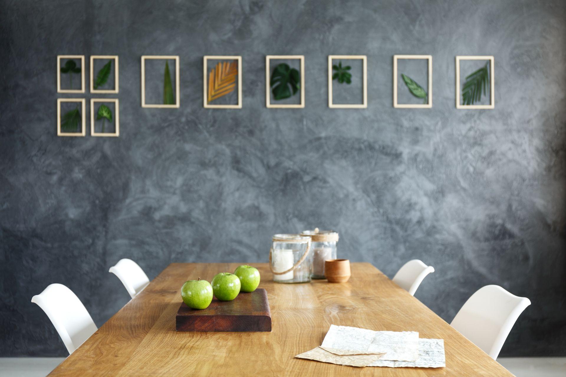 Ein minimalistischer Essbereich mit einem Esstisch aus Holz, drei grünen Äpfeln als Dekoration und eleganter Wandgestaltung mit Blättern in Bilderrahmen.