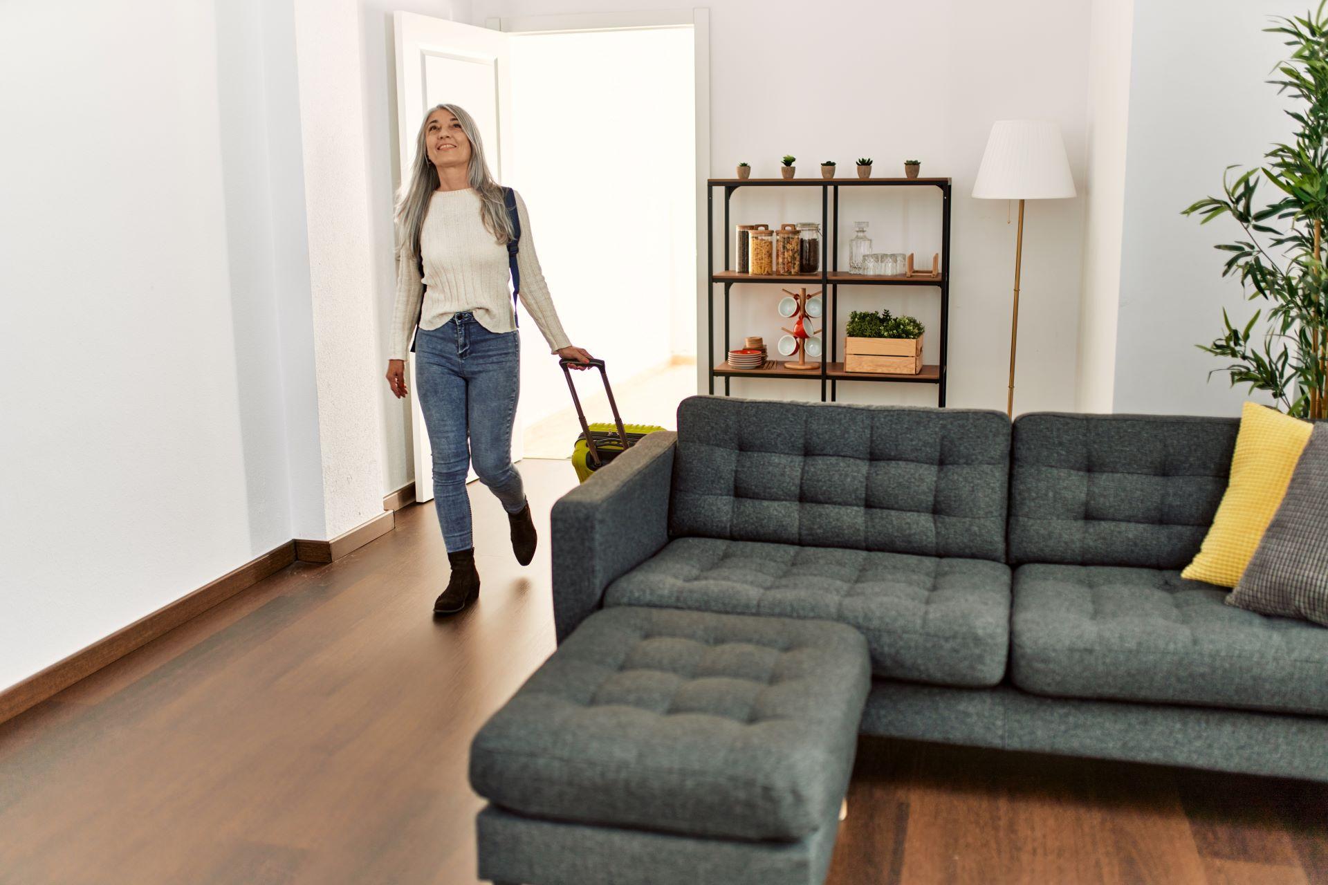 Ankunft in der Ferienwohnung – Eine Frau mit grauen Haaren und Rucksack betritt lächelnd eine modern eingerichtete Ferienwohnung mit gemütlichem Sofa, stilvollen Regalen und heller Atmosphäre.