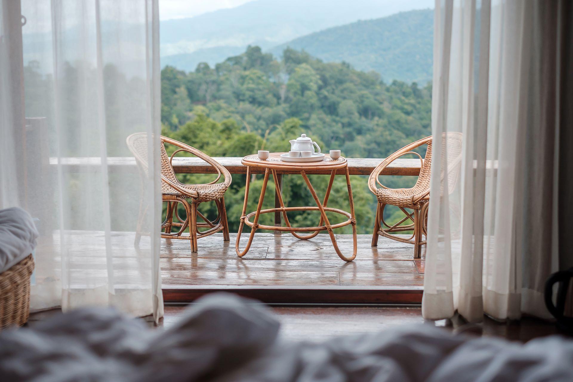 Gemütliche Terrasse mit Bergblick – Ein einladender Balkon mit Rattanmöbeln, gedecktem Teetisch und atemberaubender Aussicht auf bewaldete Hügel, gesehen durch geöffnete Vorhänge.